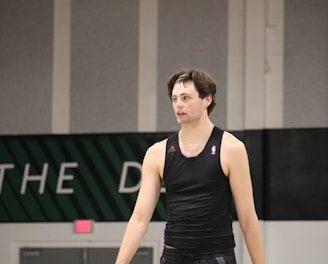 A person wearing a sleeveless black sports top is standing indoors in a gymnasium. The background includes a gray wall with green and white lettering partially visible.